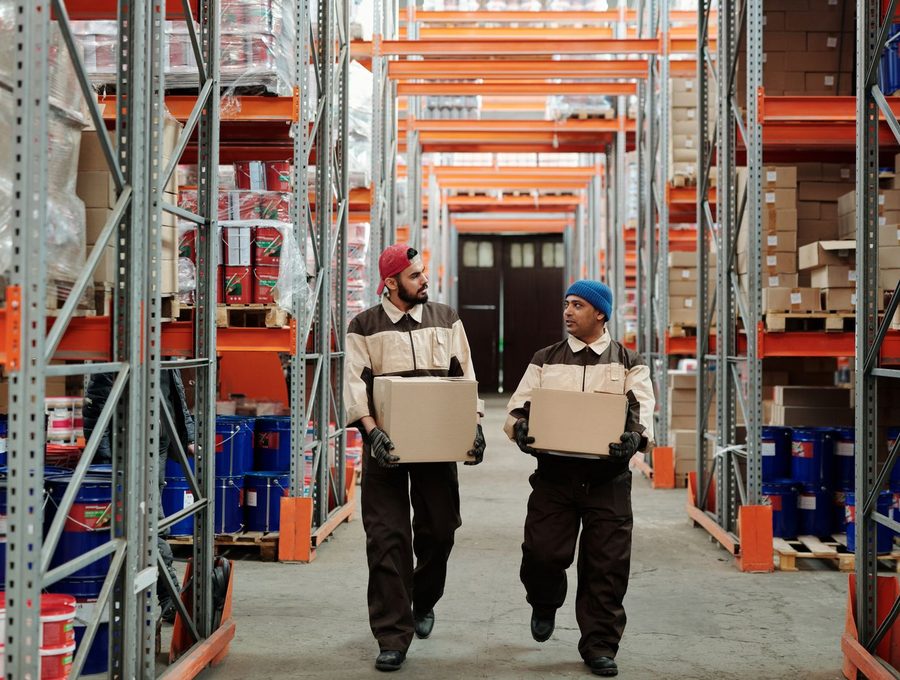 Two warehouse workers carrying boxes down a racking aisle